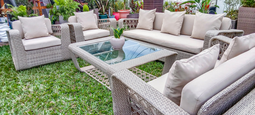 Outdoor patio furniture set with woven gray wicker-style chairs, a sofa, and a glass-top coffee table on green grass, with beige cushions and potted plants in the background.