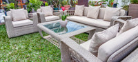 Outdoor patio furniture set with woven gray wicker-style chairs, a sofa, and a glass-top coffee table on green grass, with beige cushions and potted plants in the background.