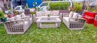 A 5-piece outdoor patio furniture set made of woven wicker, featuring a 3-seater sofa, 2-seater armchairs, a loveseat, and a rectangular coffee table with a glass top, arranged on a green lawn.