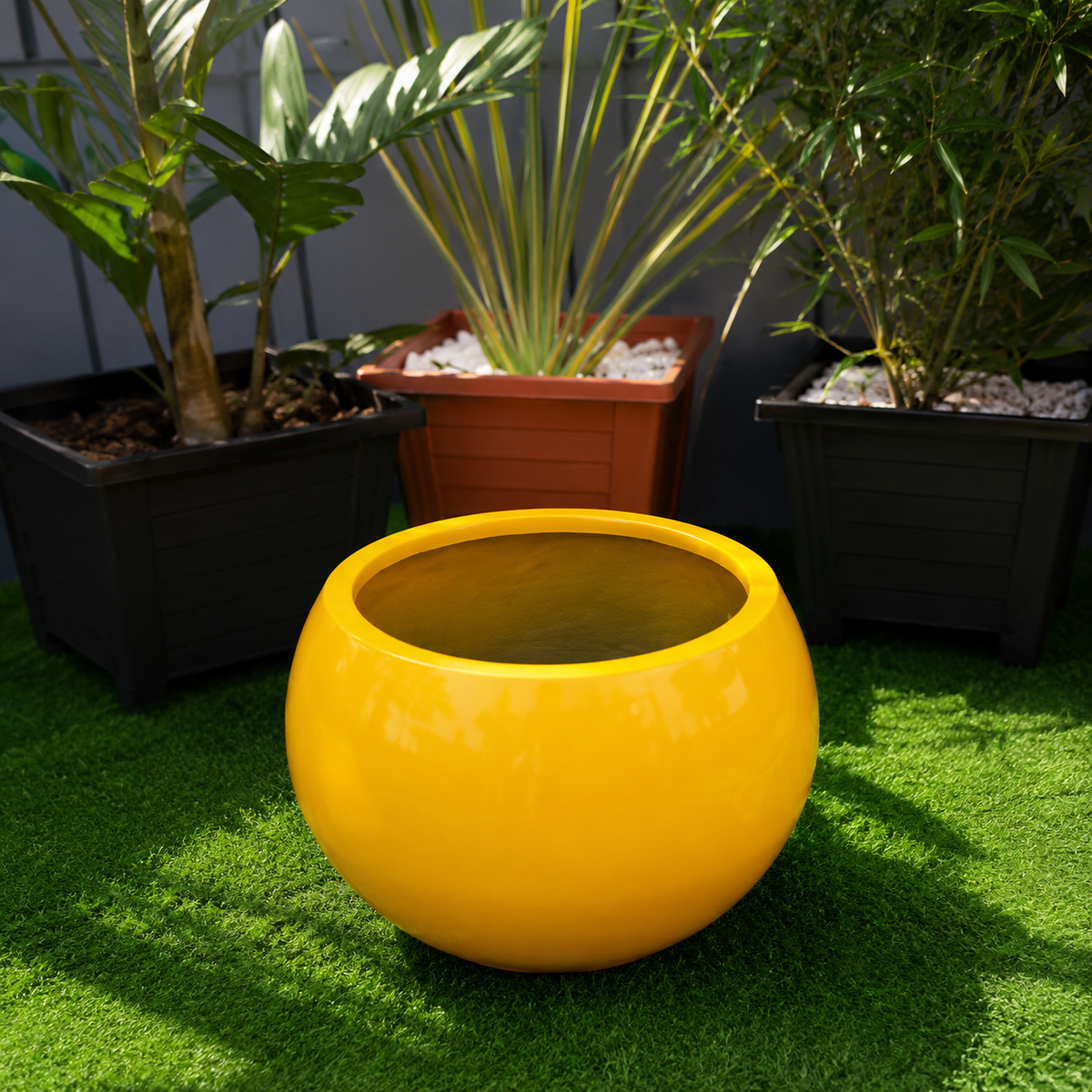 A glossy, spherical yellow planter pot filled with water, placed on green artificial grass in front of potted plants.